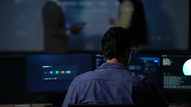 A seated developer framed from behind holds still at his monitors while the pair beyond him keep working through the architecture inside a dark AI startup office.