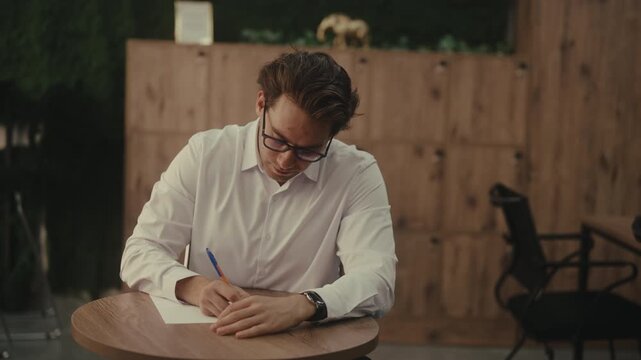 Dedicated professional male in white shirt and glasses sitting at table, writing on sheet of paper, pausing to reflect before continuing task with renewed focus on modern office room, slow motion.