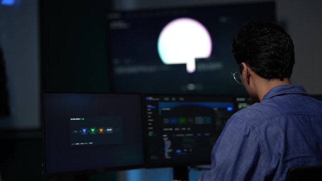 A curly haired engineer alone at a desk leans into his monitors in a dark room, the only light a lamp and the code on the screen at an AI company in Bangkok, Thailand.