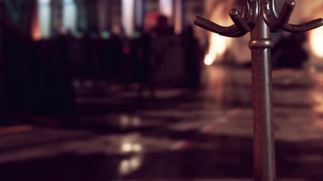 The setting shows a street at night with reflections on the ground. A metal stand is in the foreground, with blurred people and lights further back. The atmosphere is lively and busy.