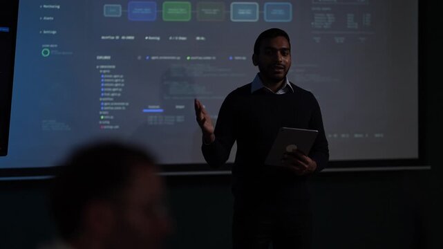 A speaker in a dark knit sweater presents an AI agent architecture to a seated audience, the huge screen behind him full of live metrics inside a dark AI startup office.