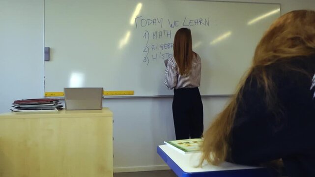 Young female teacher writing the day's learning subjects on a whiteboard while a student watches attentively from her desk. Education and learning concept in a modern classroom setting