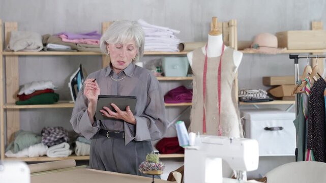 Attentive old tailoress drawing dress design in tablet in sewing workshop . High quality 4k footage
