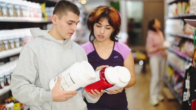 Uncertain young female and male customers reviewing jars of protein in sports food shop 