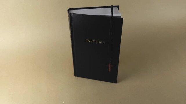 Holy bible with a black leather cover and a crucifix necklace bookmark slowly rotating on a light brown background, representing Christian faith, scripture study, and spiritual guidance