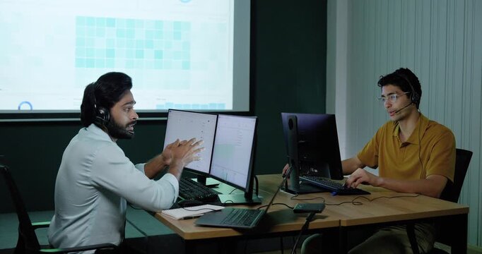 Two adult male technology professionals discuss software information across computer workstations, gesturing while collaborating on system analysis, digital workflow and business planning in office.