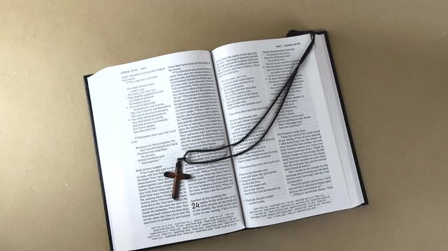 Open holy bible lying on a light brown surface with a wooden cross necklace placed on the pages, symbolizing faith, prayer, and christian devotion in a still life representation of spirituality