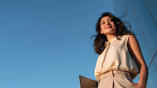 Professional businesswoman confidently looking up to the clear sky, celebrating career success, future vision, and aspiration with a laptop outside a modern office building