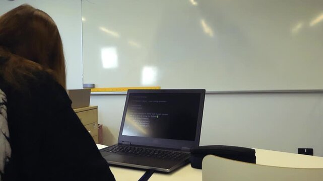 Young female student sitting at a desk in a classroom while learning to code on a laptop, with a command line interface program visible on the screen and a whiteboard in the background