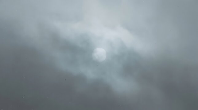 A bright sun glowing behind a thick layer of soft grey clouds in the overcast sky