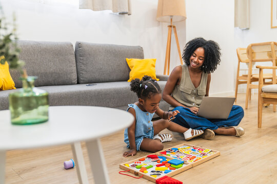 African american mother using a laptop while her young daughter plays with a wooden puzzle on the living room floor at home