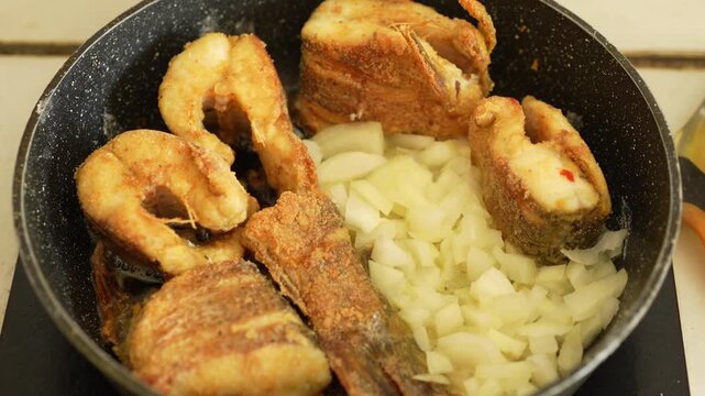 A top-down shot of golden fried fish pieces being cooked in a pan alongside a large portion of chopped white onions.