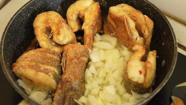 A top-down shot of golden fried fish pieces being cooked in a pan alongside a large portion of chopped white onions.