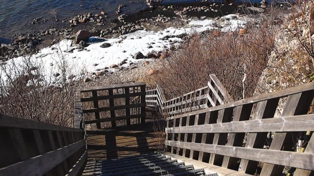 High angle view descending a long wooden staircase down a cliff to the rocky shore of the Baltic Sea, where patches of snow remain on the beach during a sunny early spring day in Paldiski