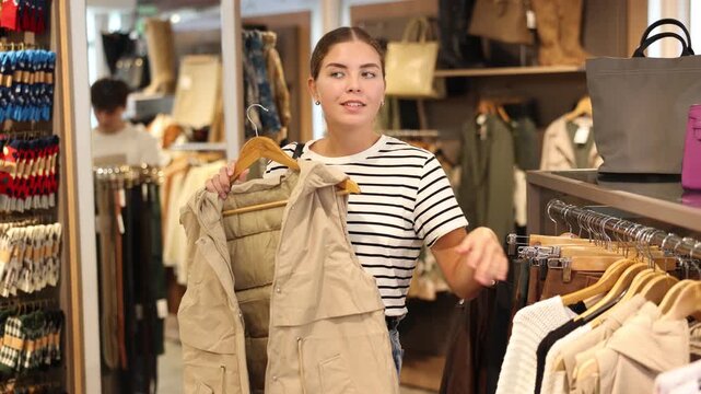 After inspection of stores assortment, girl chooses vest on hangers and closely examines its details, feels texture of fabric. Buyer thoughtfully goes through folds touch of sleeveless jacket fabric 
