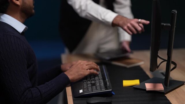 A close view of hands at a keyboard keeps typing while a standing colleague leans across the desk to point something out inside a dimly lit machine learning startup office.