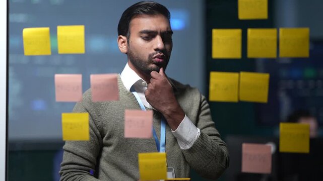 A thoughtful engineer at a transparent board runs a hand along his jaw before reaching up to move one card in the workflow inside a dimly lit machine learning startup office.