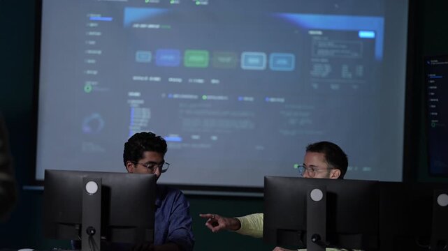 A wide view of a darkened engineering floor shows two developers at their desks dwarfed by an enormous projected agent dashboard at an AI company in Bangkok, Thailand.