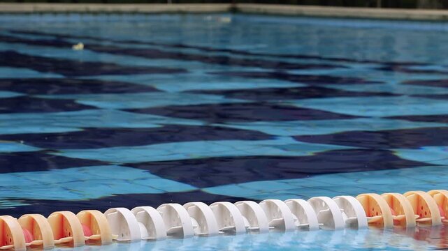 Swimming pool lane divider floats on water surface. Orange and white plastic floats form a line. Blue water ripples gently around the divider. Ideal for fitness, sports, or aquatic activity scenes