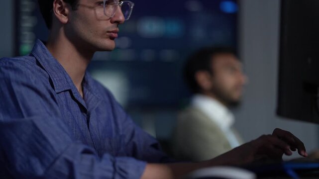 A developer late in a long shift presses his fingers to his eyes while a colleague keeps working further down the room inside a dimly lit machine learning startup office.
