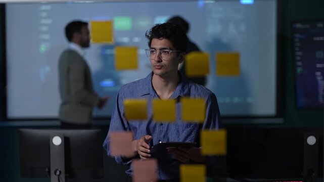 An engineer with a tablet at the note wall looks up from his screen and smiles as the answer to the sequence finally lands inside a dimly lit machine learning startup office.
