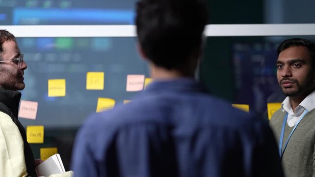 A developer seen from behind at the board faces two colleagues through the notes as one of them offers a page from his notebook inside a dark AI startup office.