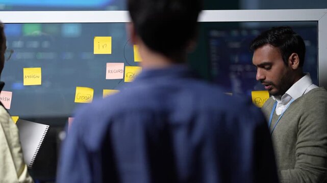 A team writing at a glass planning wall keeps moving, one engineer noting a decision down while another reaches up to the notes inside a dark AI startup office.