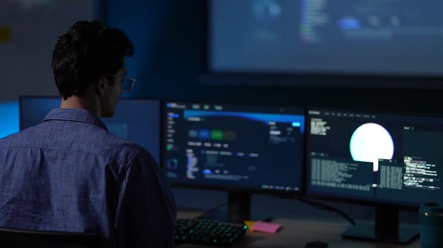 A young engineer in half silhouette keeps his eyes on the monitors as a small lamp throws a bloom of light across the desk inside a dimly lit machine learning startup office.