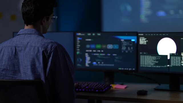 A developer working a late shift alone scans between two monitors of telemetry with the rest of the office dark behind him at an AI company in Bangkok, Thailand.