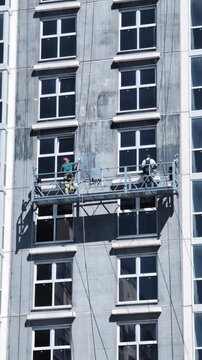 Construction workers on elevated suspended platform scaffold applying primer coat to exterior facade of high-rise building wall for preparation of painting activity. Vertical shot