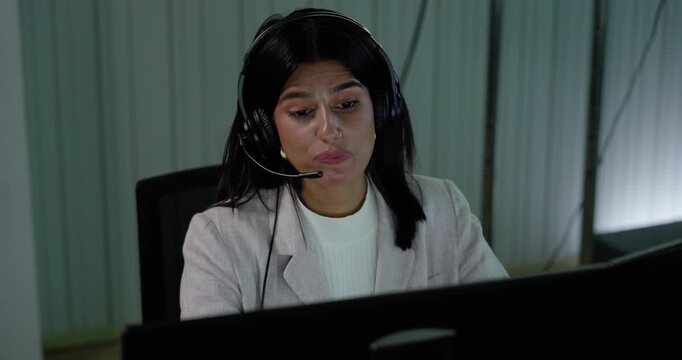 Adult female customer support professional wears headset, speaks toward computer monitor and changes facial expression while handling digital communication during call center service work in office.