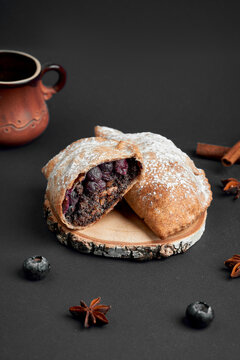 Halved sweet baked pie with poppy seed and blueberry filling dusted with powdered sugar on wood slice. Dessert pastry on dark background.