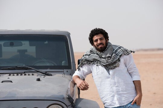 Relaxed confident Arab man wearing shemagh scarf leans against dusty off road SUV smiling in vast open sandy desert ideal for MENA lifestyle travel