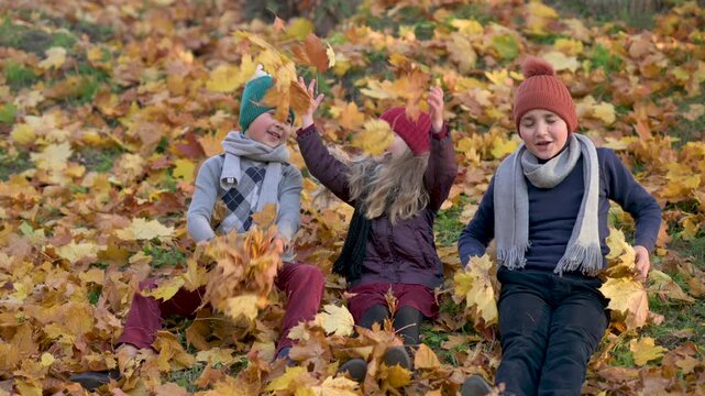 hree happy kids, two boys and a girl, playing together in autumn park, picking up and throwing colorful fallen leaves. Landscape orientation.