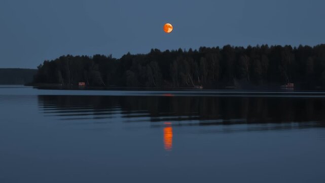 Orange full moon glowing brightly above lake and dark forest line, casting vibrant reflection upon still water of a tranquil lake surface at dusk twilight evening. Picnic camping