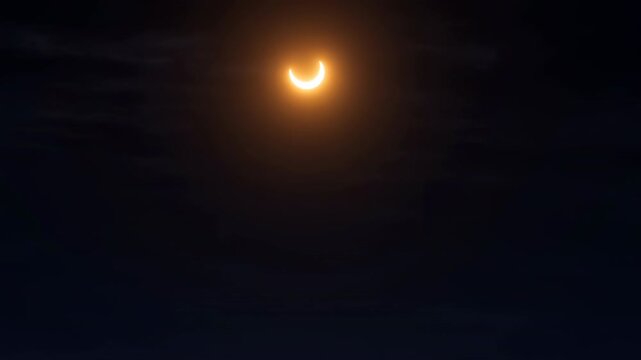 Crescent sun glows through drifting dark clouds during a partial solar eclipse, surrounded by warm orange light and deep shadow in a centered view of the dramatic sky