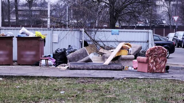 Large pile discarded household furniture miscellaneous garbage bags urban sidewalk beside overflowing trash bins, depicting neglect environmental concern waste management problem.