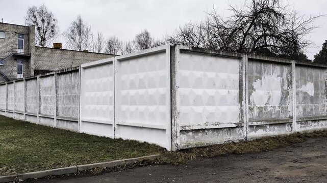 Long weathered concrete fence sections with geometric patterns and peeling white paint line property boundary next to dry grass strip, grey building, and bare trees under overcast sky