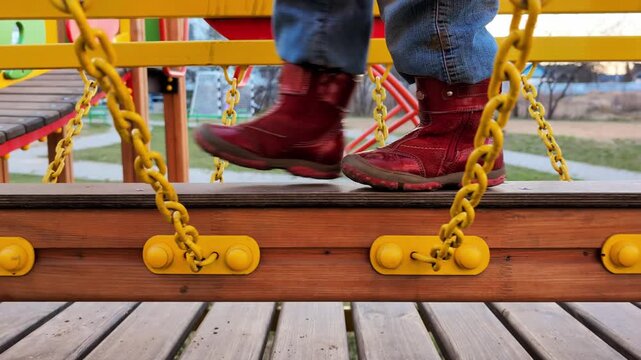 Young person feet in red boots walking on wooden playground bridge with yellow chains, outdoor activity, childhood fun and development, safe play area structure