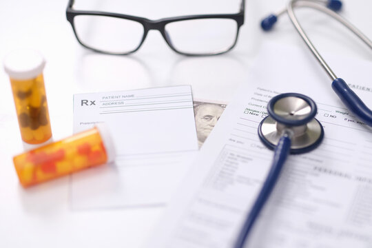 Bright medical workspace with prescription forms, stethoscope, eyeglasses, pill bottles and partial cash note on white desk, illustrating healthcare, medication review, patient paperwork and bribery