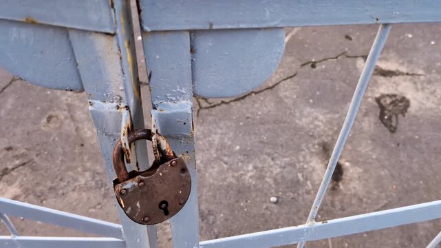 Rusty padlock secures old metal gate, showing signs of wear and corrosion, positioned outdoors over cracked concrete surface with focus on intricate locking mechanism details.