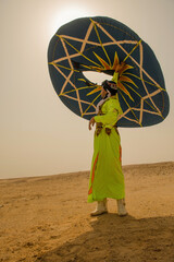 Colorful folk performer in ornate embroidered robe raises a massive geometric teal disc high overhead on sunlit Sahara desert sands ideal for cultural © .shock