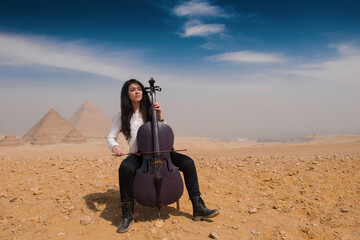 Young female classical musician playing cello on the Giza plateau with iconic ancient pyramids and wide blue sky behind her ideal for arts travel and © .shock