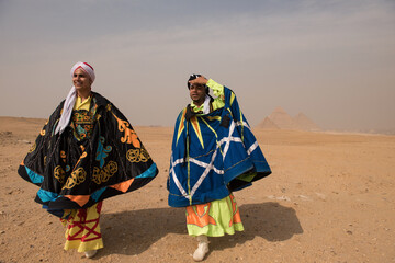 Two Egyptian tanoura folk performers wearing vivid ornate capes and turbans pose candidly before the iconic Great Pyramids of Giza on the desert © .shock