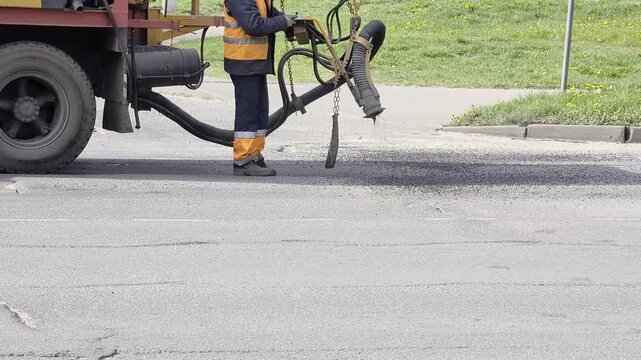 Worker in safety vest applies asphalt mixture to damaged road surface with specialized road repair machine, performing essential street maintenance outdoors during day, copy space