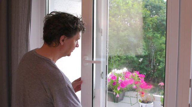 A man washes the windows in his apartment. He uses a spray bottle to clean the glass. Flowers can be seen outside the window. This is part of his regular cleaning tasks