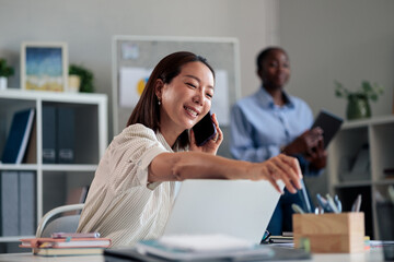 Fototapeta premium Asian businesswoman smiling and talking on smartphone in office
