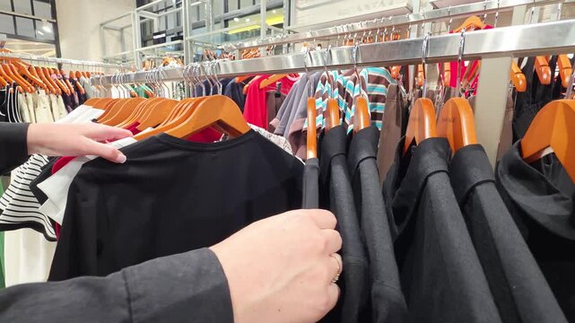 Person hands browsing through clothing items hanging on a rack in a retail store, selecting garments for purchase from a wide array of fashion choices.