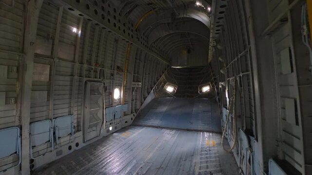Interior cargo aircraft fuselage empty. Metal walls, exposed framework, and bare floor. Ramp visible at rear, some windows providing light. Industrial aircraft cabin, large transport plane inside view
