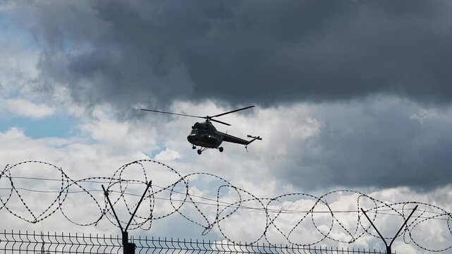 Military helicopter flies over razor wire fence on cloudy day, dark clouds loom in sky, air patrol over secure restricted area, aerial transport security